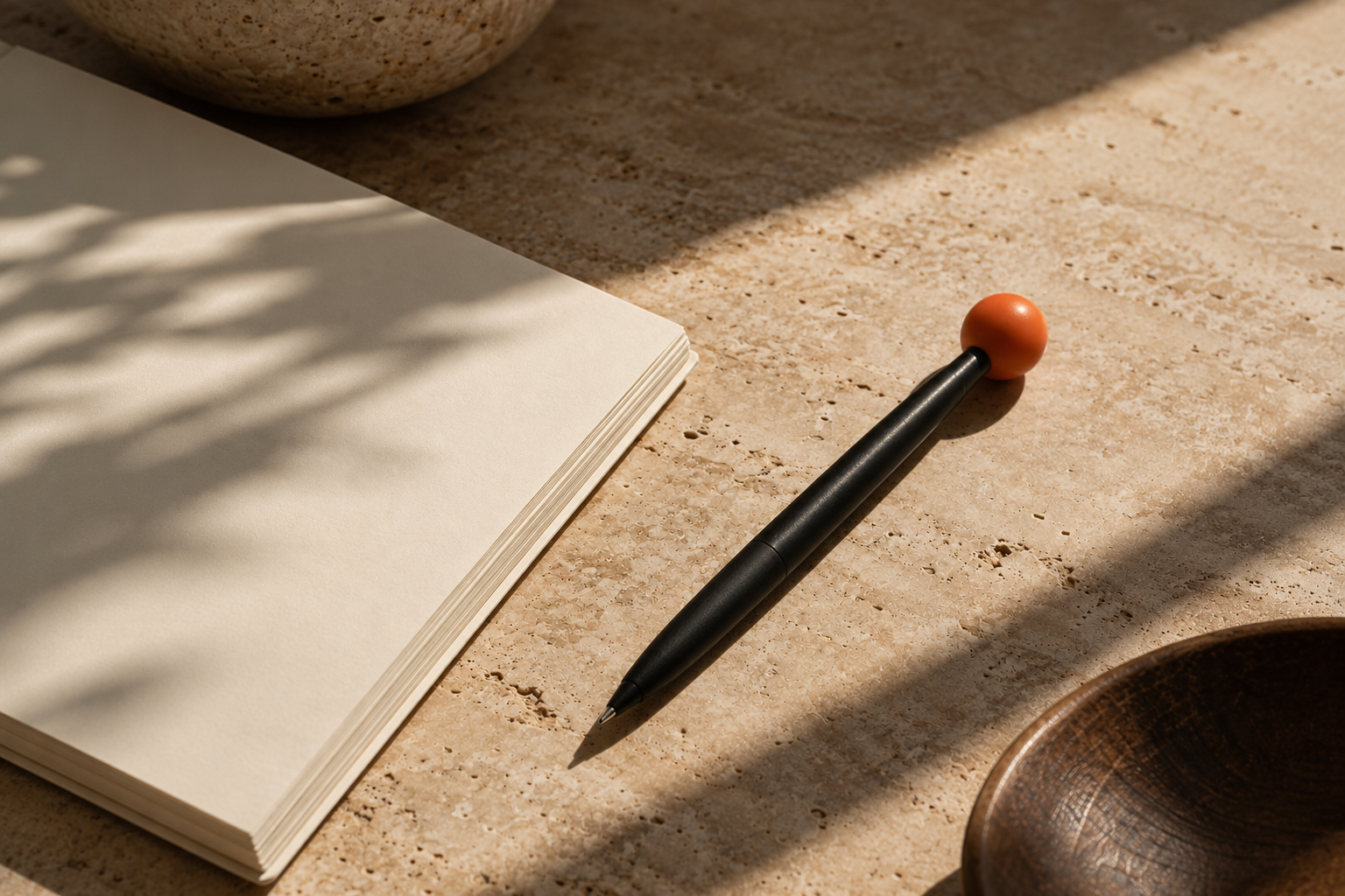 A cream notebook beside a black pen with a bright orange ball-shaped top, lying on a warm travertine surface in sunlight